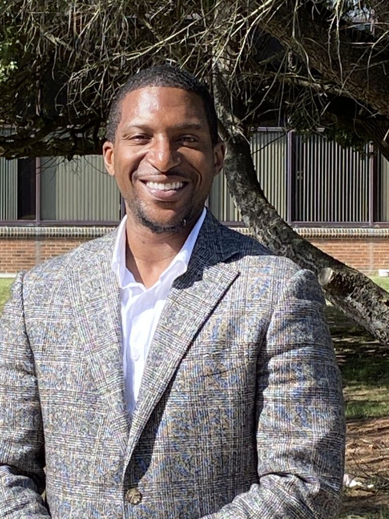 Dr. Lloyd M. Talley, smiling in a light grey suit jacket with an open-collar white shirt
